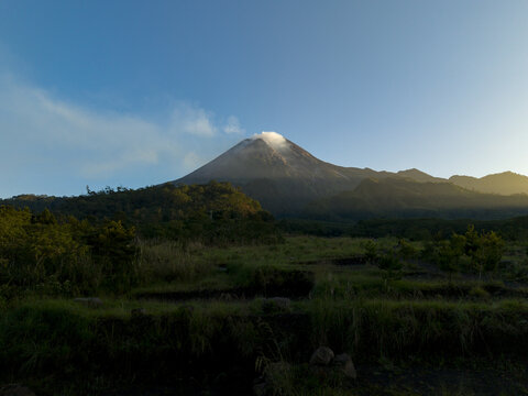 Aerial view of Bunker Kaliadem Merapi and the smoking peak of Mount Merapi volcano surrounded by lush green forests under a clear sky in Kecamatan Pakem, Daerah Istimewa Yogya, Indonesia.