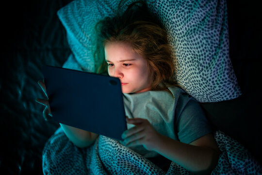 Kid facing tablet light in bed at night. Teenager lying in bed watching tablet at night.