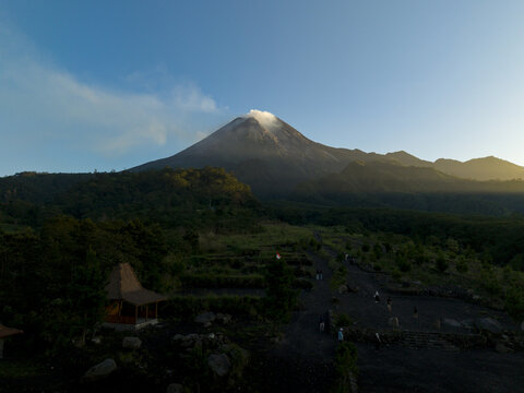 Aerial view of Bunker Kaliadem Merapi with the smoking peak of Mount Merapi surrounded by lush green forests under a clear blue sky Kecamatan Pakem, Daerah Istimewa Yogya, Indonesia.