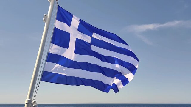 View of the Greek national flag billowing in the sea breeze on a ferry crossing the Aegean Sea, set against a clear summer-blue sky.