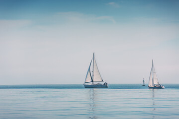 Naklejka premium Two Sailboats With White Sails and With Crew Aboard Sailing on a Calm Foggy Sea Under a Pale Blue Sky Near the Horizon