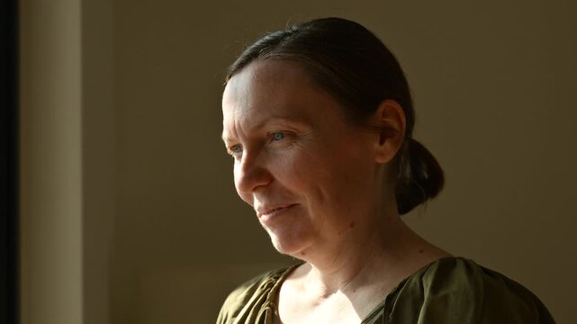 Thoughtful middle-aged woman standing by a window in warm natural light, looking outside with a serious contemplative expression, quiet interior portrait. 4K