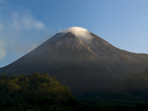 Aerial view of Mount Merapi volcano with steam rising from the crater above lush tropical forests under a clear blue sky Kecamatan Pakem, Daerah Istimewa Yogya, Indonesia.