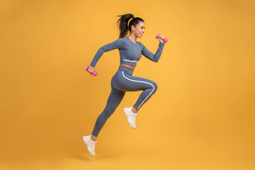 Fototapeta premium Energetic young woman in blue activewear jumping while holding pink dumbbells on a yellow background, representing fitness motivation and dynamic strength training 