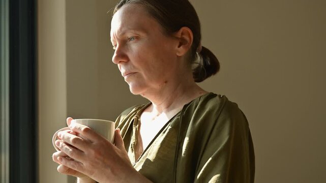 Thoughtful middle-aged woman holding cup of coffee or tea by window in warm morning light, quiet moment of reflection at home. 4K