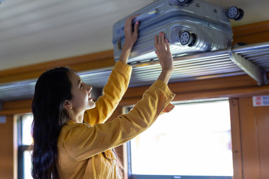 Young woman on a train stowing a suitcase in the overhead rack, smiling as she prepares for a long journey and enjoys the freedom of public transit travel.
