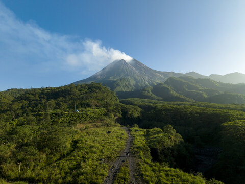 Aerial view of Bunker Kaliadem Merapi and the smoking peak of Mount Merapi surrounded by lush green forests in Kecamatan Pakem, Daerah Istimewa Yogya, Indonesia.