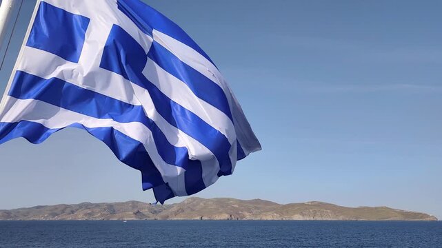 View of the Greek national flag billowing in the sea breeze on a ferry crossing the Aegean Sea, set against a clear summer-blue sky.