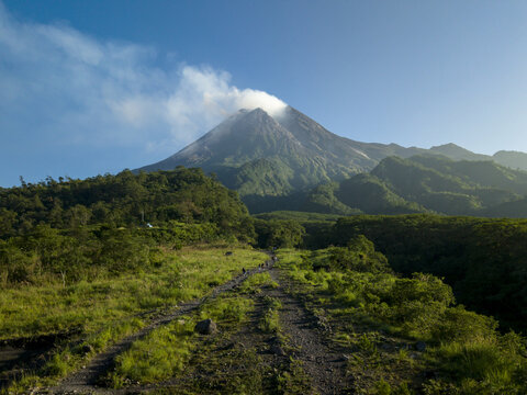 Aerial view of Bunker Kaliadem Merapi and the smoking crater of Mount Merapi volcano with lush green slopes and a dirt path Kecamatan Pakem, Daerah Istimewa Yogya, Indonesia.