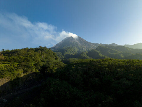 Aerial view of Mount Merapi with smoke rising from the crater above lush green forests and hills under a clear blue sky in Kecamatan Pakem, Daerah Istimewa Yogya, Indonesia.