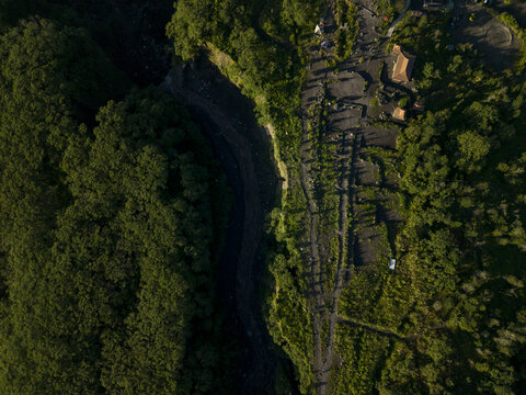 Aerial view of Bunker Kaliadem Merapi area featuring a dark volcanic riverbed surrounded by lush green forest and small structures in Kecamatan Pakem, Daerah Istimewa Yogya, Indonesia.
