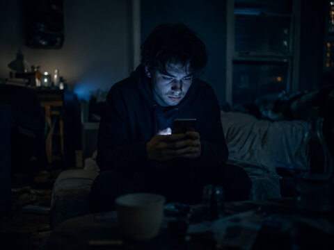Young man scrolls on smartphone alone in a dark bedroom, moody blue cinematic lighting, ideal for digital wellbeing, social media and tech marketing ads.