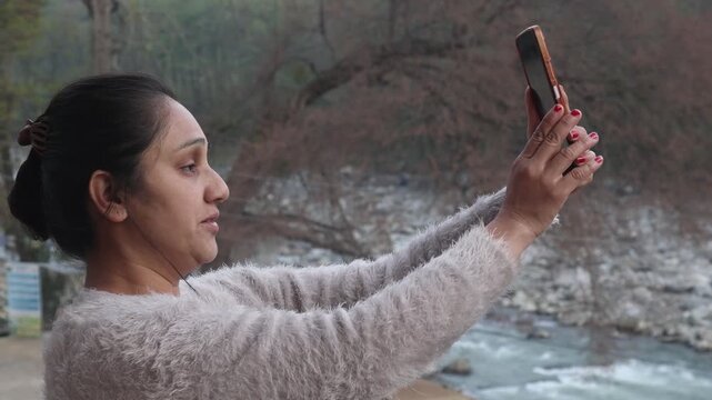Indian woman taking a selfie with a mobile phone while enjoying a peaceful mountain river view in the Himalayas India. Natural outdoor lifestyle scene showing travel, happiness, and rural mountain bea