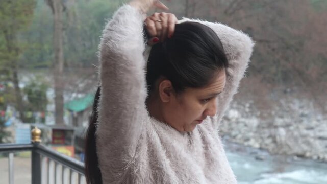 Indian woman tying up her hair while standing in a peaceful Himalayan mountain environment in India. Natural outdoor lifestyle scene with beautiful river valley and rural mountain landscape in the bac