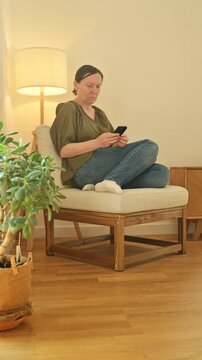 Middle-aged woman sitting and using smartphone, focused or concerned expression in warm cozy home lighting, 4K vertical video