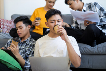 Fototapeta premium Four Young Asian Men Study Together in Cozy Living Room, Drinking Coffee and Using Laptop
