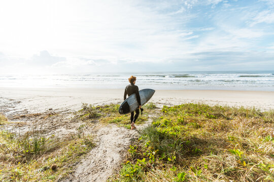 Surfer in wetsuit walking down a sandy track with surfboard at beach