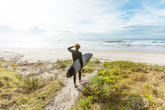 Surfer in wetsuit walking down a sandy track with surfboard at beach
