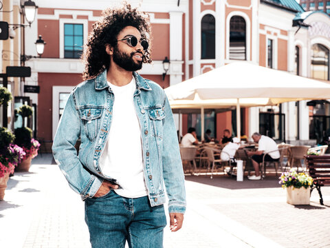 Handsome smiling hipster  model.Sexy unshaven Arabian man dressed in summer jeans jacket clothes. Fashion male with long curly hairstyle posing in street at sunset. Cheerful and happy. In sunglasses
