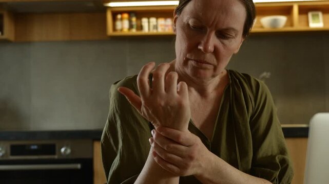 Mature woman massaging sore wrist near laptop at home workspace, discomfort from computer work and repetitive strain lifestyle concept. 4K.