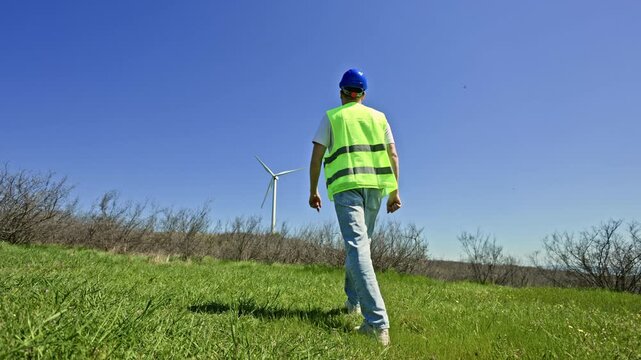 A windmill engineer walks on a green field under a clear blue sky. The engineer approaches a wind turbine during a site inspection for maintenance and project planning.