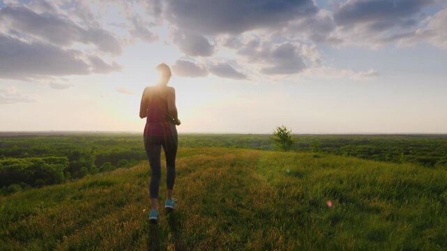 Woman running on grassy hill at sunset with beautiful clouds and forest in background.