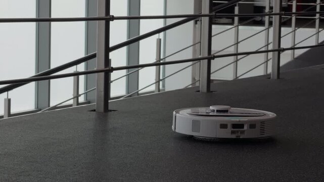 Wireless robotic vacuum maneuvering along walkway beside metallic balustrade with modern windows