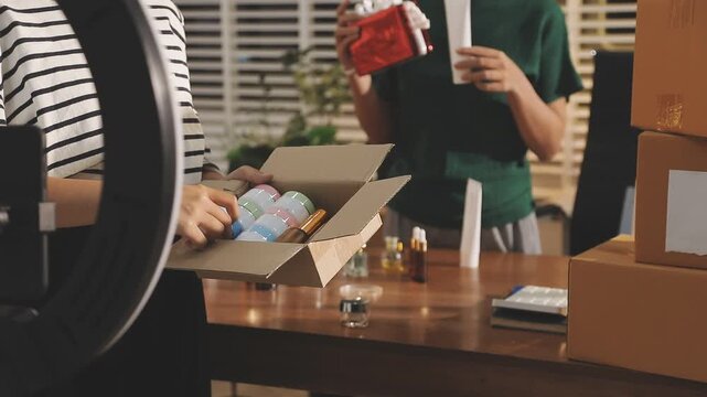 Young beautiful Asian girl reviewing cosmetic and skin care product with new collection's promotion selling on social media online live blurred phone screen surrounded beauty merchandise. Stratagem.