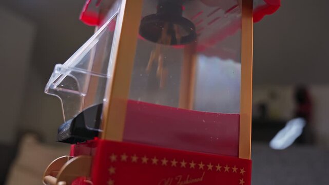 unpopped corn kernels falling inside a vintage red popcorn machine. Warm indoor lighting highlights the old fashioned aesthetic during home movie night preparation.