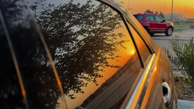 a vibrant golden sunset and tree silhouette reflected on a clean car window. Cinematic evening lighting in an outdoor parking setting with shallow depth of field.