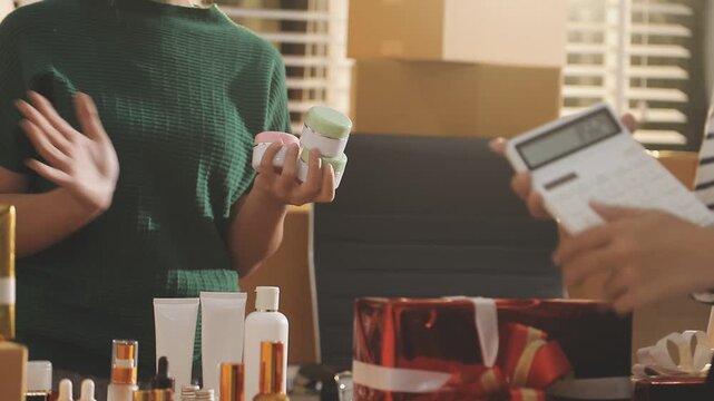 Young beautiful Asian girl reviewing cosmetic and skin care product with new collection's promotion selling on social media online live blurred phone screen surrounded beauty merchandise. Stratagem.