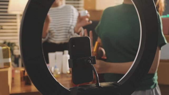 Young beautiful Asian girl reviewing cosmetic and skin care product with new collection's promotion selling on social media online live blurred phone screen surrounded beauty merchandise. Stratagem.