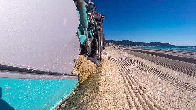 Extreme Close-up of Beach Cleaning Machine Leveling Sand, Side View Tracking Shot, Sandy Shore Maintenance with Sea Background. The background shows a vast sandy beach, deep blue sea with gentle waves