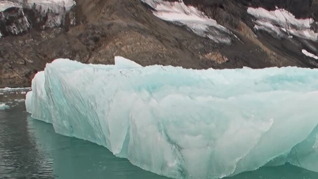 Explore the Arctic Ocean's serene yet formidable environment. A magnificent blue iceberg drifts past snow-dusted mountains and icy waters, captured during an extreme polar expedition.