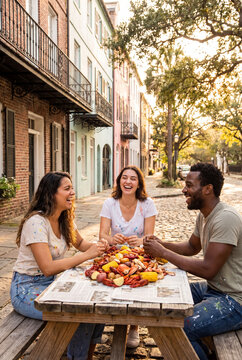 Three diverse friends laugh together while eating a delicious seafood boil with corn and crab on a historic cobblestone city street during sunset.