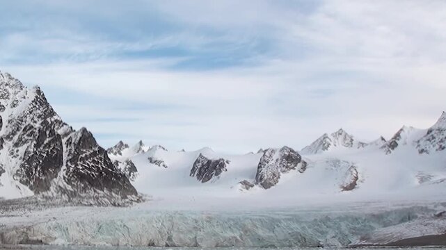 Witness the colossal grandeur of an Arctic glacier, its shimmering ice face meeting tranquil waters. Majestic snow-capped mountains loom under a serene daytime sky.