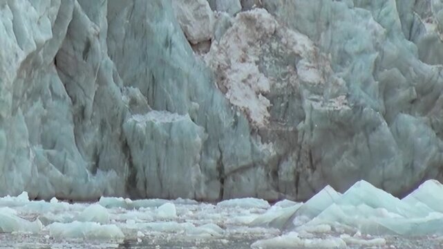 Experience a colossal glacier wall in the Arctic. Its pristine crystalline surface rises majestically above tranquil waters, reflecting shimmering ice fragments under a bright sky.