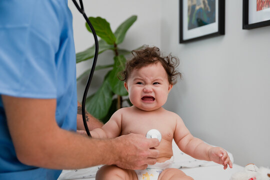 Infant sitting upright, looking at the camera and crying while the doctor is listening to her heart with a stethoscope, with copy space