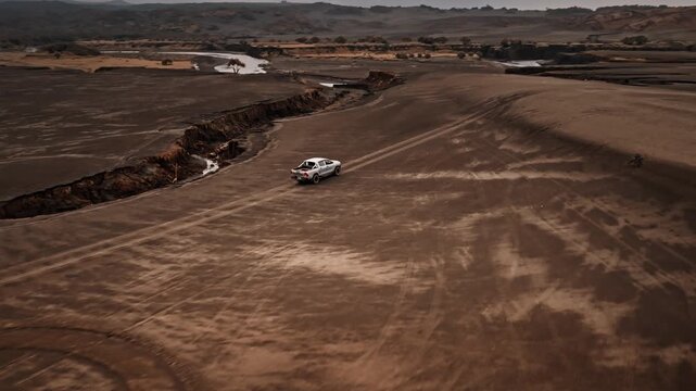 Pickup truck car driving along tire tracks on barren ash plain next to dry riverbed, depicting arduous off road adventure and exploration in a remote volcanic landscape. Aerial view drone flight