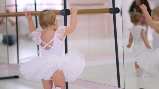 Little happy girl in white ballerina dress standing at barre near mirror in dance studio, a childhood dream concept