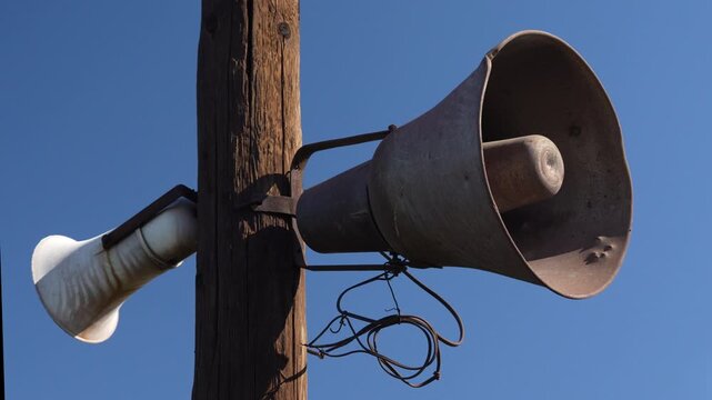 Slow panning shot of old metal loudspeaker mounted on wooden pole against blue sky. Concept of public announcement system, alert and emergency communication.