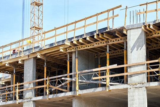 Multi-story building under construction, featuring concrete pillars, wooden formwork for floor slabs, and temporary yellow support props beneath a clear blue sky