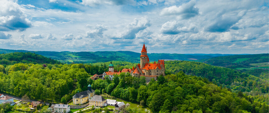 Aerial view of Bouzov Castle with its iconic red-roofed towers nestled among dense green forests and rolling hills under a cloudy sky in Bouzov, Olomoucky kraj, Czechia.
