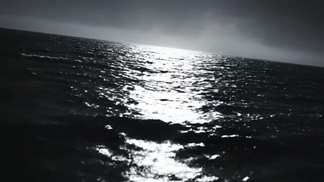 Monochrome footage of a dark ocean horizon with sparkling sunlight reflecting on the rippling water, evoking a sense of calm and mystery.