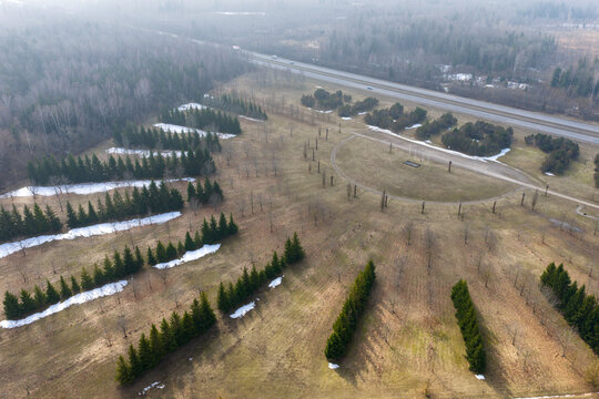Aerial view of rows of evergreen trees and a semi-circular path with patches of melting snow near a highway and misty forest in Lithuania.