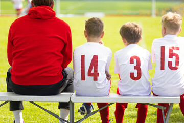 Naklejka premium Youth Soccer Coach and Substitute Players in Numbered White Jerseys Sitting on Bench Watching Game - Junior Football Team Management and Substitute