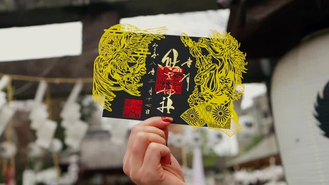 A white Latina visitor holds a black and yellow special edition goshuin. The background shows a blurry Japanese shrine.