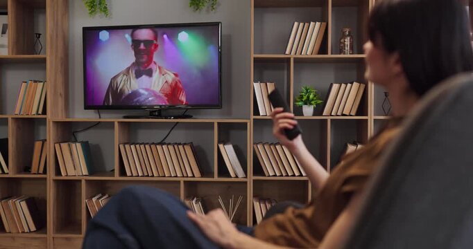 Woman watching television at home with remote. She sits on the sofa in a living room focusing on entertainment show on screen amid lights and modern lifestyle. Neat home tv leisure concept.