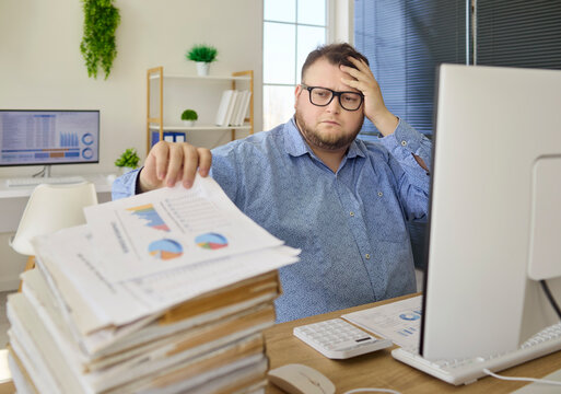 Accountant man in office facing paperwork stress. He reviews financial reports at cluttered desk, head in hand, racing a deadline and showing fatigue. Clear workplace burnout stress concept.