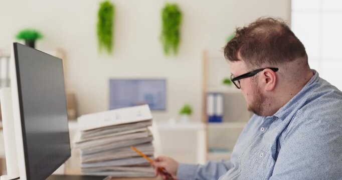 Overweight man accountant with paperwork at computer. He checks numbers by pencil near a monitor and stacks, showing focus under pressure and duties in the office. Clear concept of office focus.